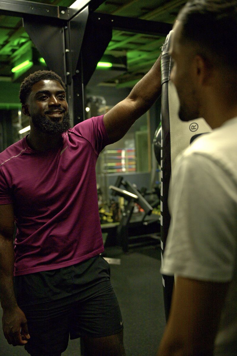 Coach Zino smiling in a magenta tee, leaning on a gym rig